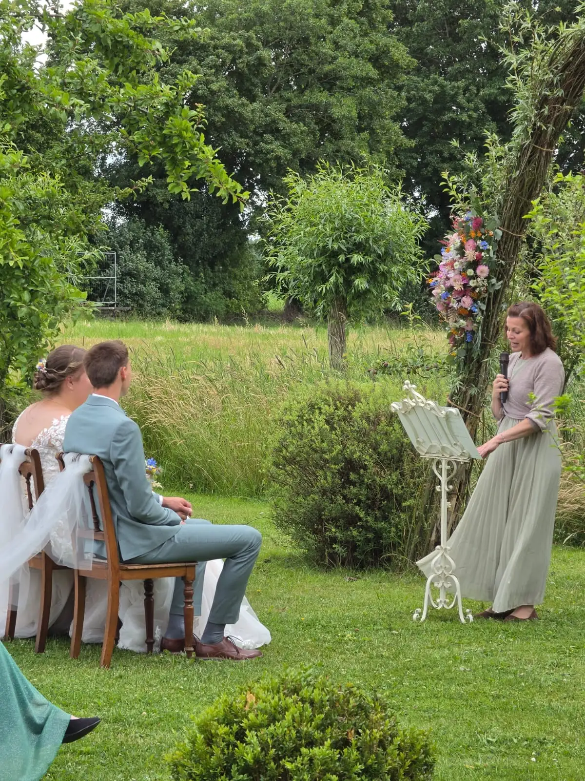 Brautpaar sitzt auf der Wiese vor der Traurednerin am Notenständer, daneben ein blumengeschmückter Bogen
